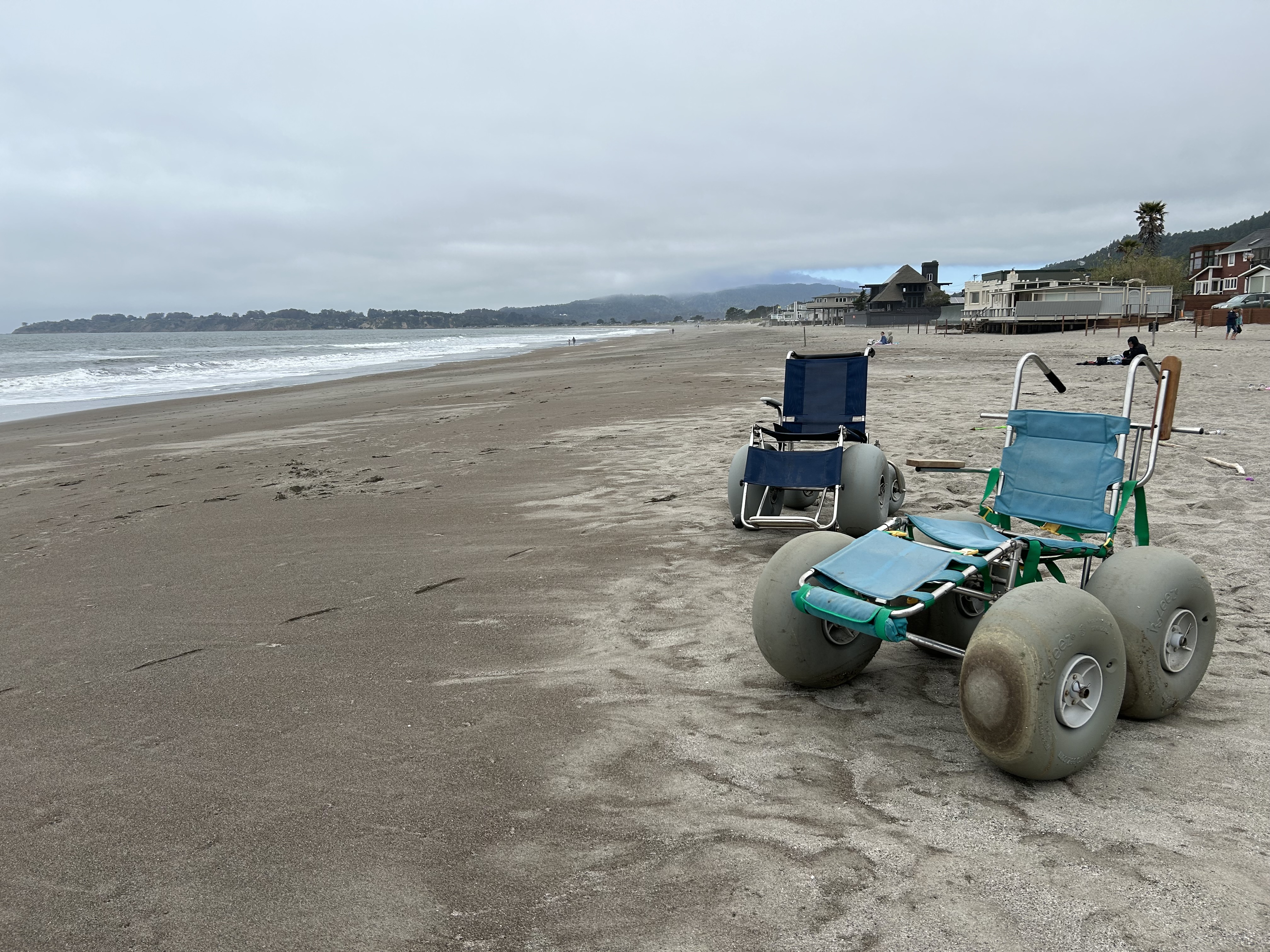 Two beach wheelchairs with bubble tires sit on a beach with the ocean visible behind